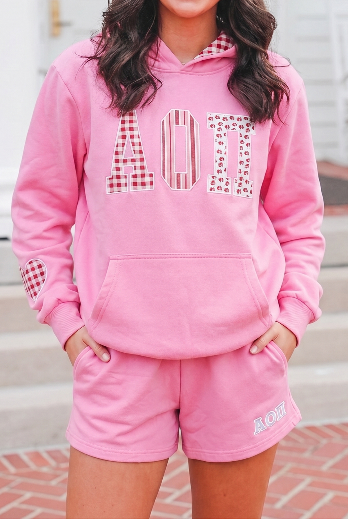 A smiling woman stands outdoors in front of a white house wearing a pink hooded sweatshirt and matching pink shorts. The hoodie has large decorative letters across the chest and pink plaid and striped pattern details on the hood lining, cuffs, and lower sleeves.